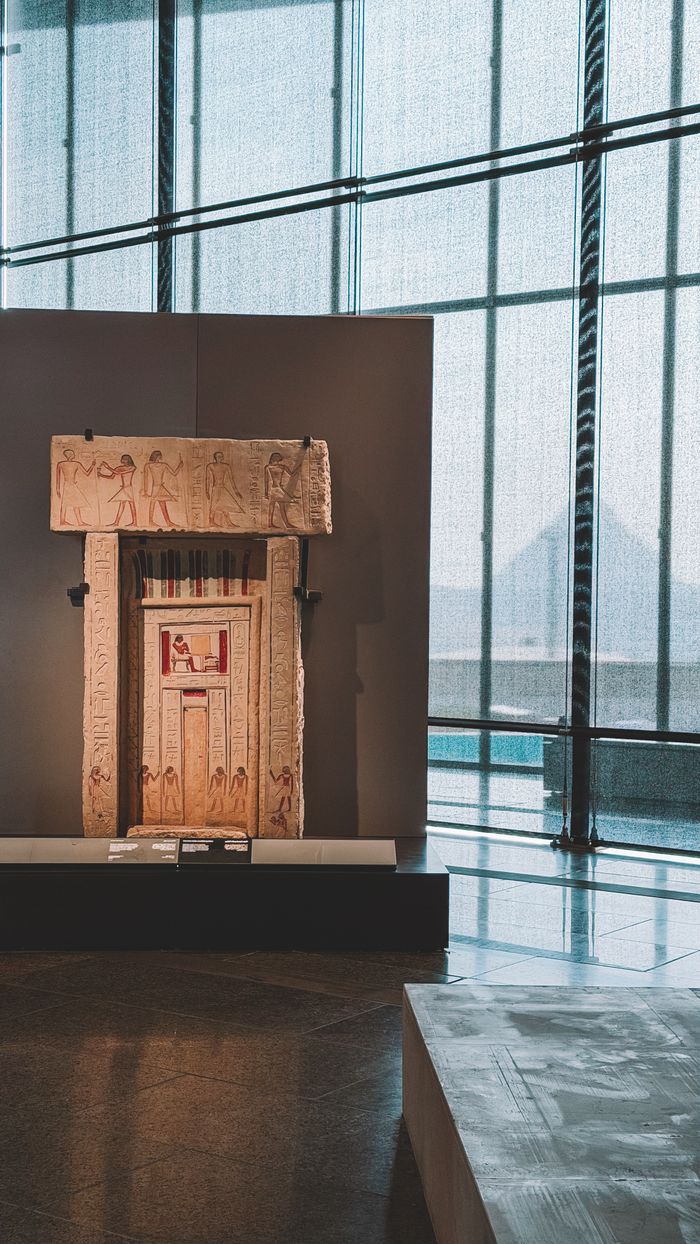 Ancient Egyptian artifact displayed in a modern museum with pyramids visible through large windows.